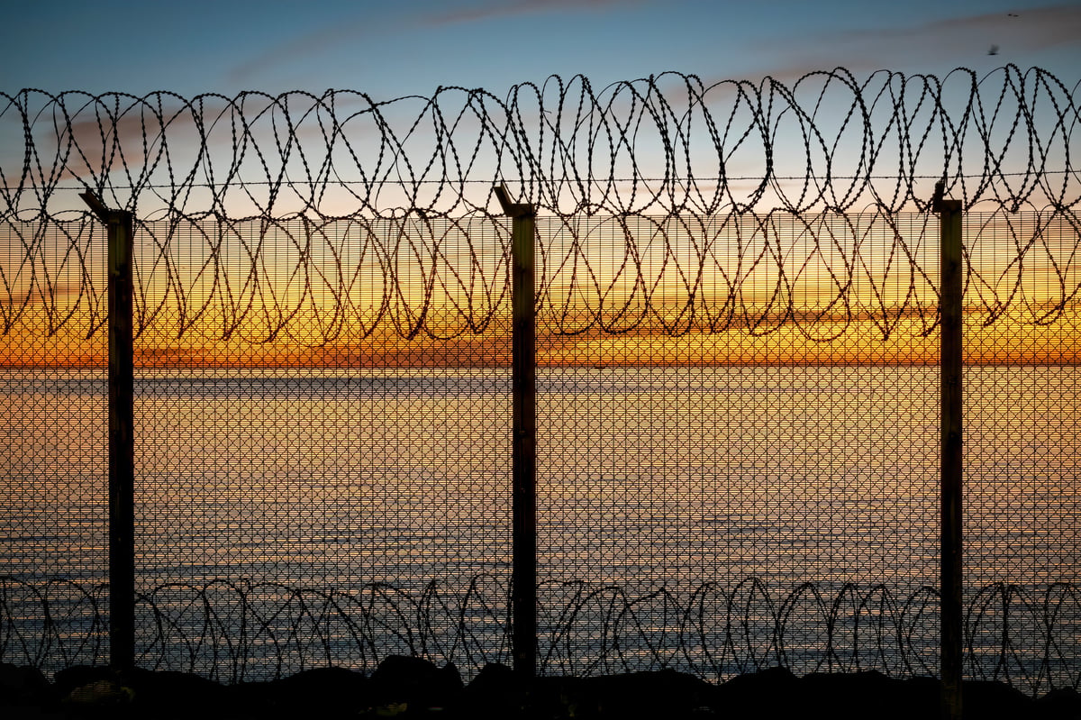 A captivating silhouette of razor wire and a chain-link fence against a vibrant sunset over the sea. Concertina wire. Security, barrier, border, freedom concept.
