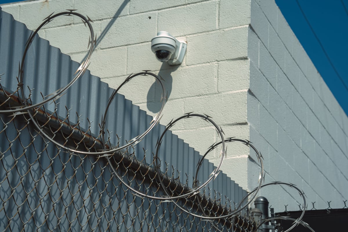 A surveillance camera is mounted on a brick wall above a chain-link fence topped with razor wire, indicating a secure perimeter
