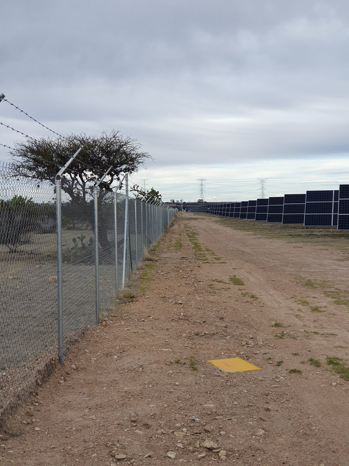Carretera de tierra con cerca de malla ciclónica y paneles solares visibles en la distancia bajo cielo nublado
