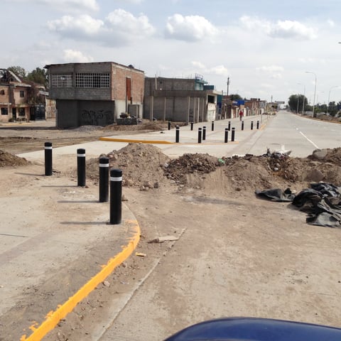 Under-construction street with black bollards, yellow curb markings, industrial buildings, and piles of dirt and materials