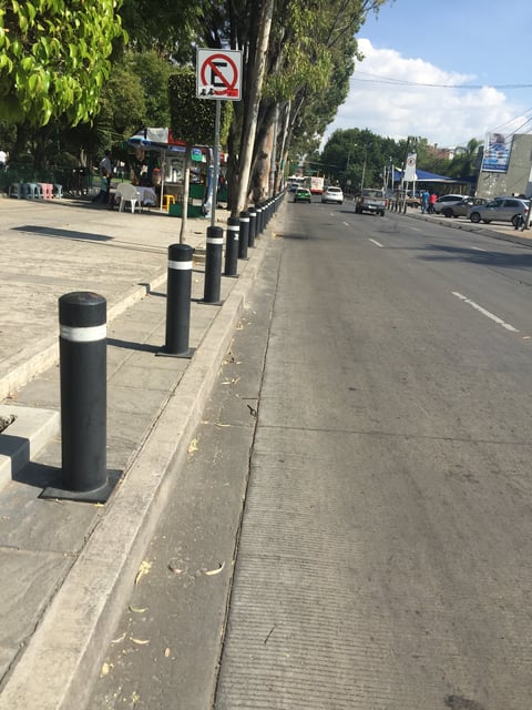 Urban street with metal bollards along sidewalk, no parking sign on pole, and parked vehicles in background