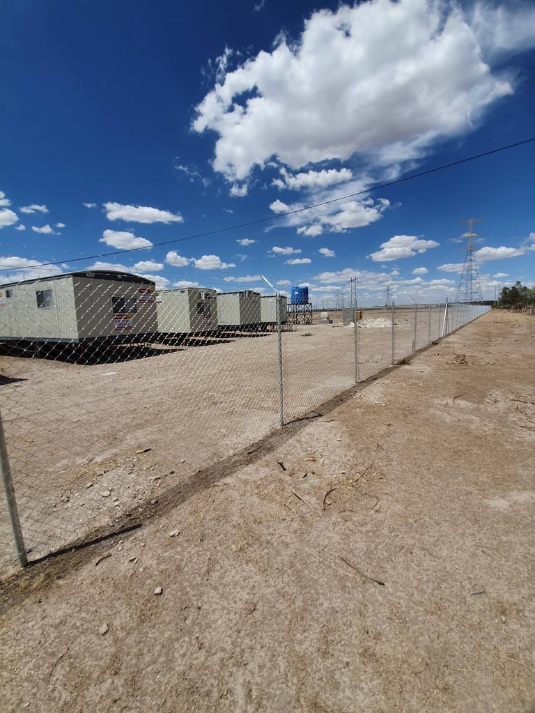Sitio industrial cercado con remolques metálicos, torre de transmisión eléctrica y cielo azul despejado