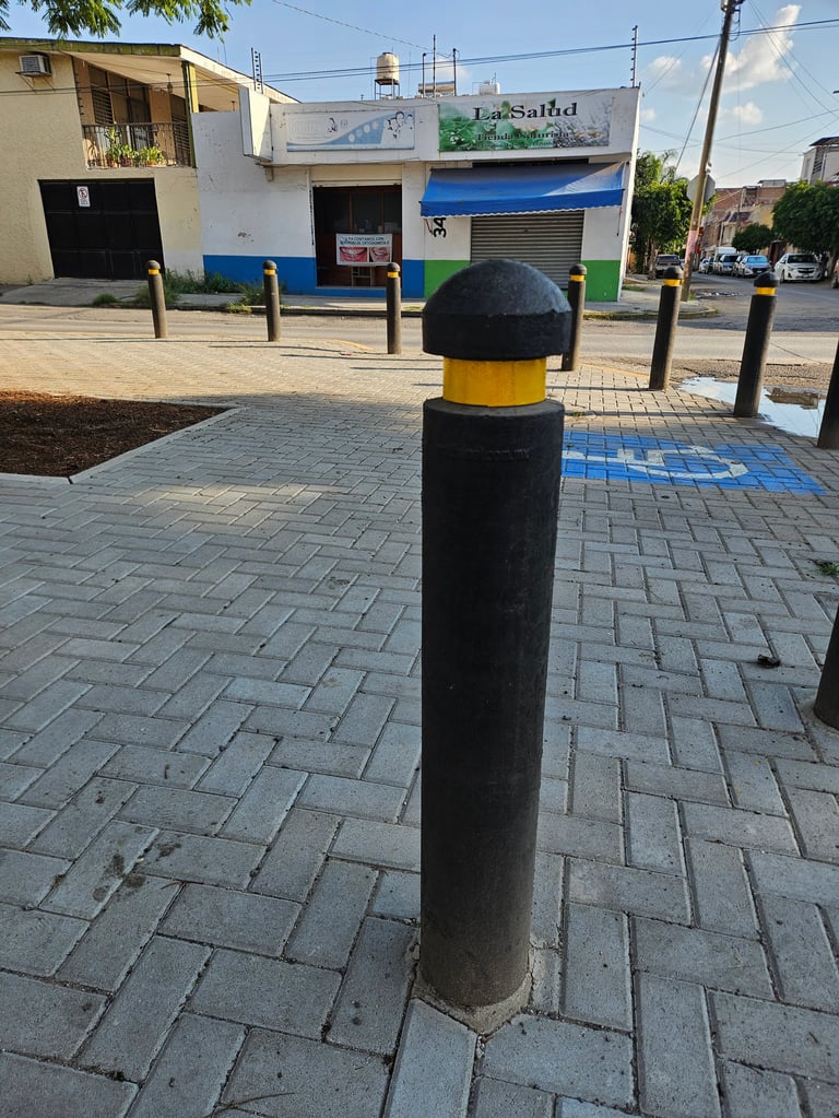 Black and yellow bollard on tiled sidewalk with storefronts and parked cars in background